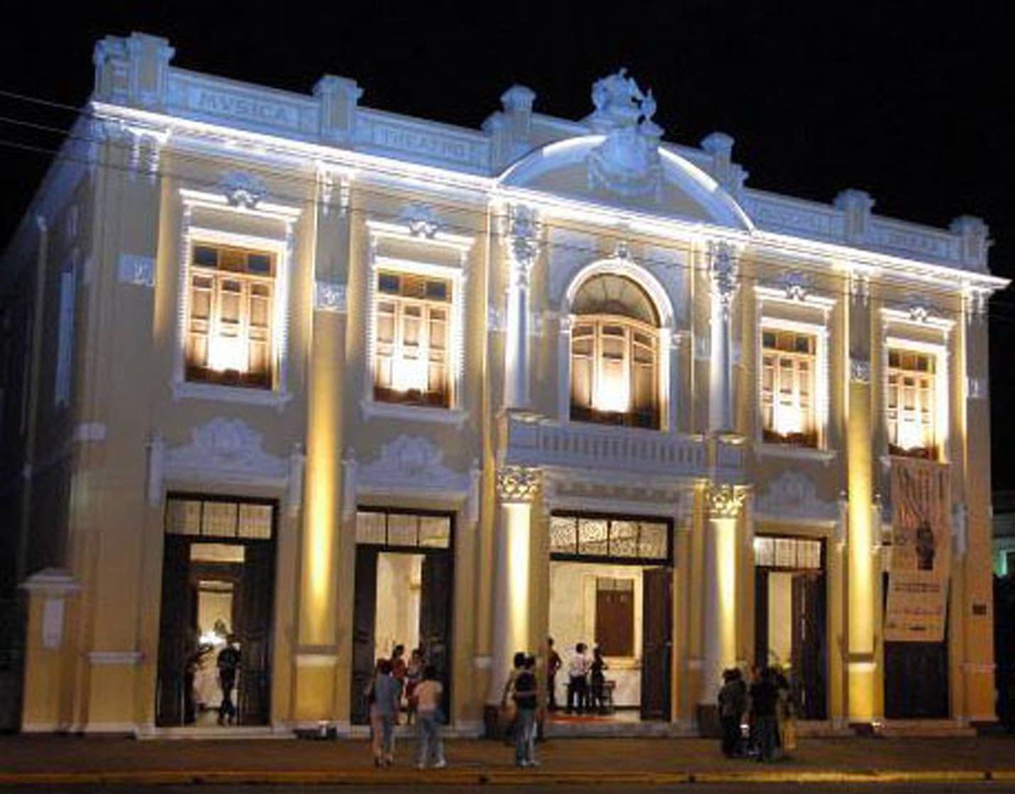 A fachada iluminada à noite, com o público na calçada.