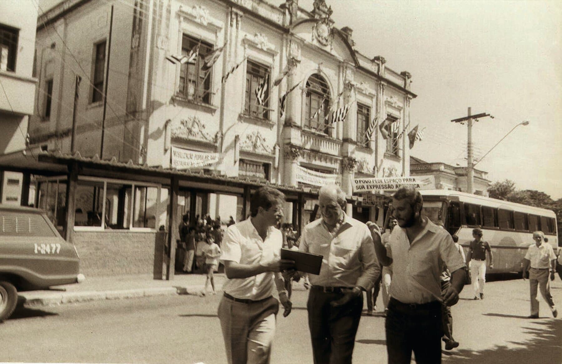 A visita do governador Franco Montoro ao Theatro, em 1986, ao lado do prefeito Sidney Beraldo: a fachada coberta de bandeiras e faixas da mobilização popular pela preservação — “O povo pede espaço para sua expressão artística”. Respaldado por mais de 2.000 assinaturas dos sanjoanenses, o município obteve a anuência do Estado, decisiva para a compra do edifício e o início do restauro.